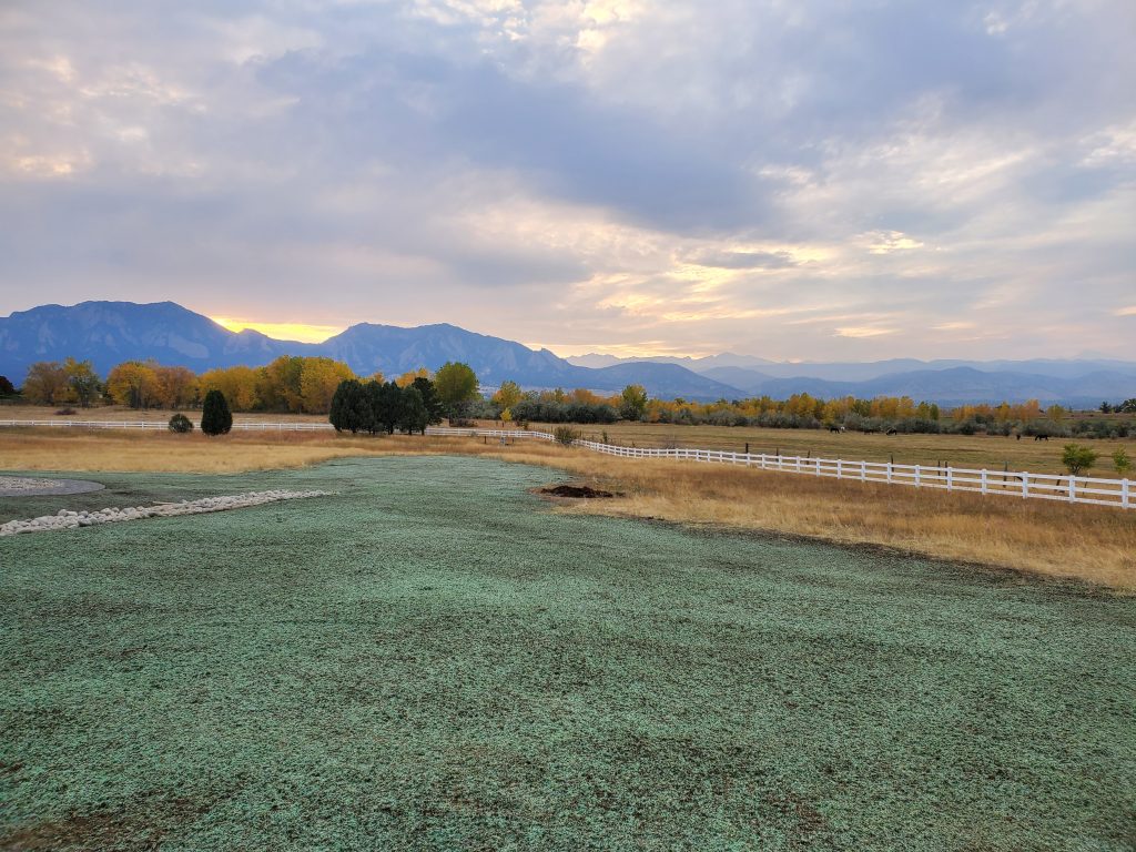 A large area of pasture inside a white picket fence with recent hydroseeding. A beautiful view of mountains and trees are in the background.